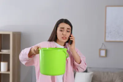 A woman holds a bucket to catch water from a leaky roof while she calls Burroughs Roofing for a repair request